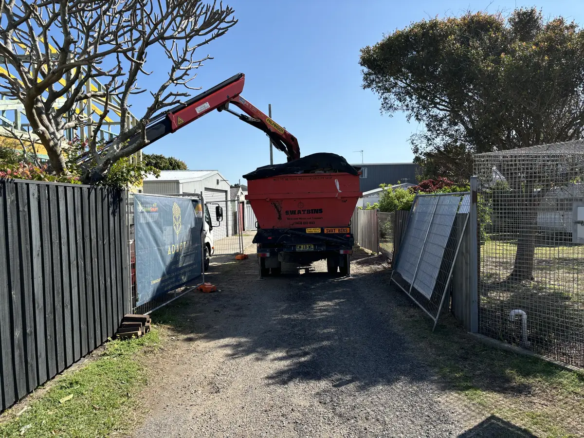 SWAT Bins crane truck lifting a skip bin down a narrow gravel lane between two fences