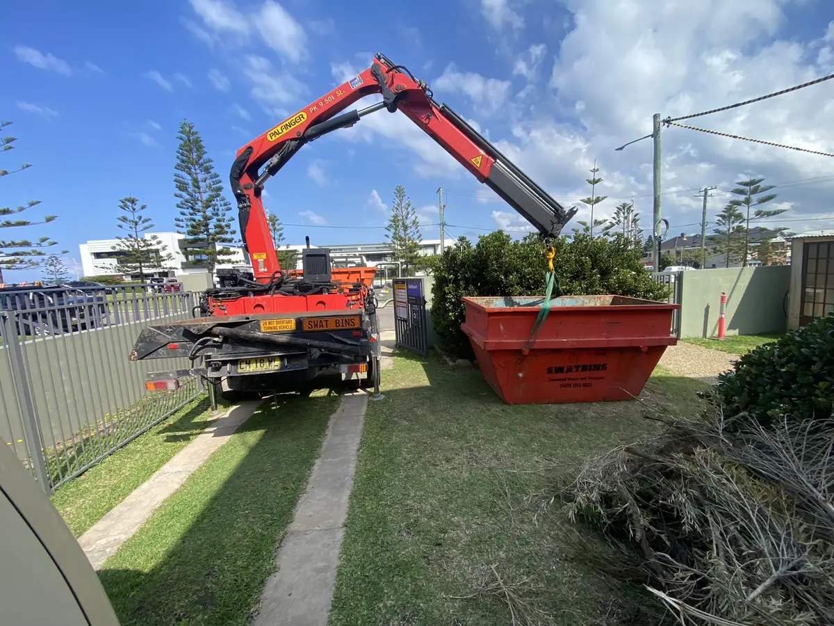 SWAT Bins crane lowering a skip bin onto a front lawn next to a pile of garden waste