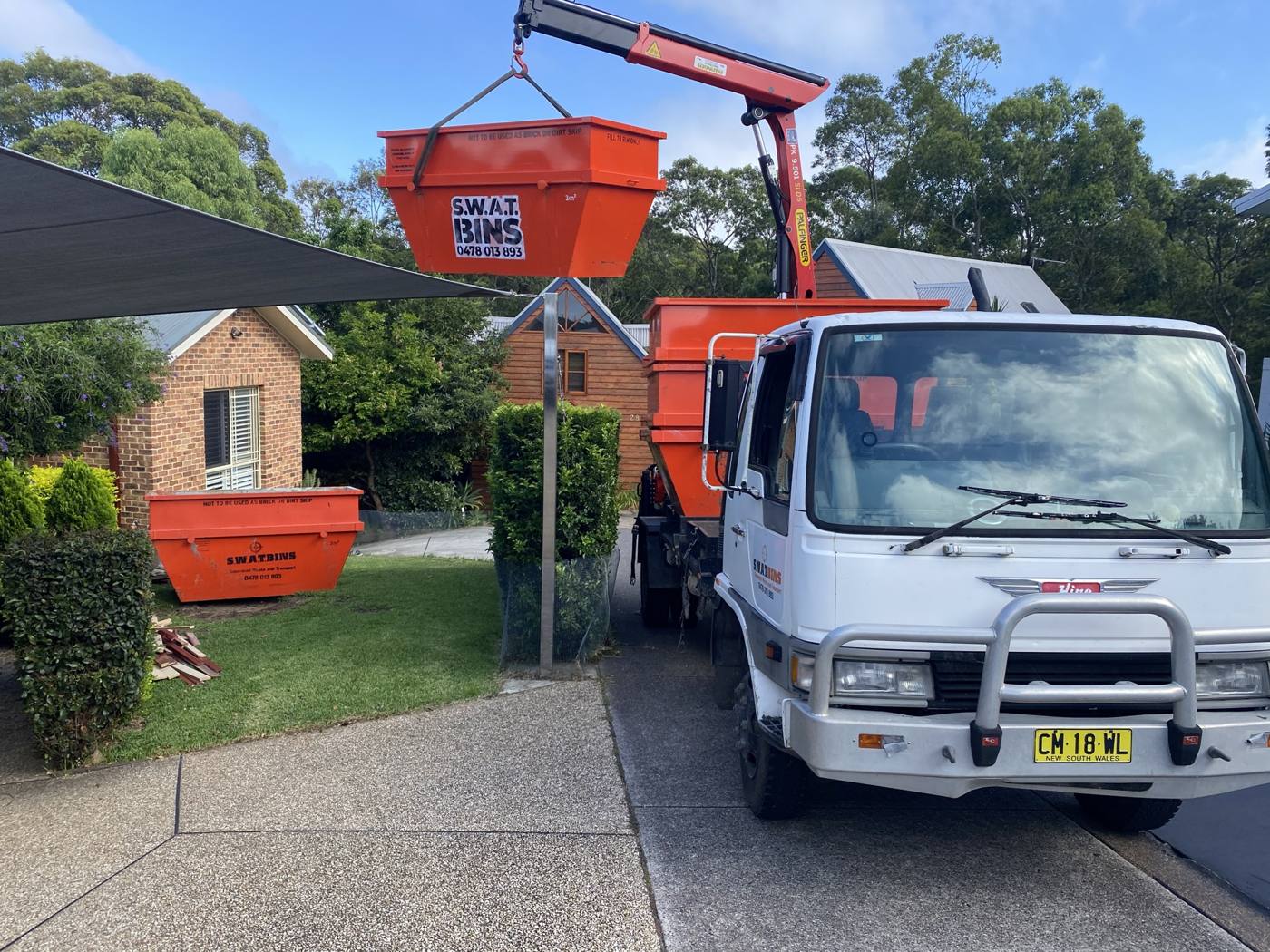 A SWAT Bins crane truck lifting an orange skip bin in a suburban driveway, with a second bin already on the front lawn