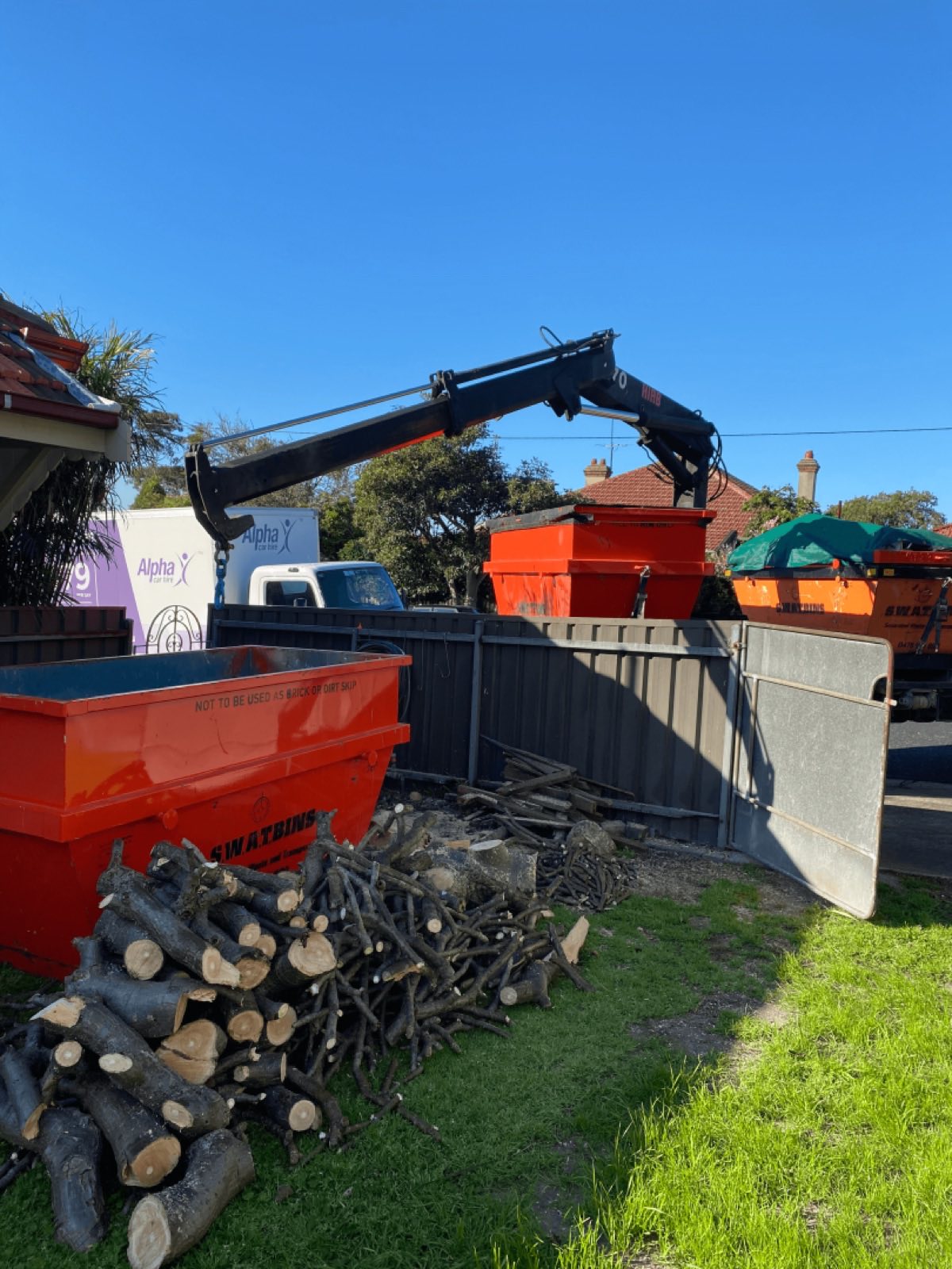 The SWAT Bins crane reaching over a backyard fence to place a skip bin beside a pile of cut branches